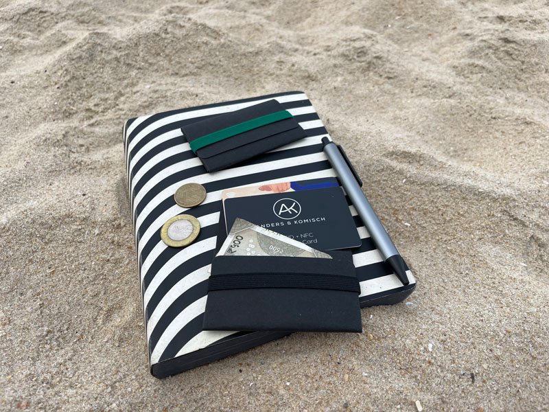 Das A&K TINY travel wallet liegt geöffnet mit Karten, Scheinen, Münzen und einer RFID Blocker Karte auf einem Journal im Sand